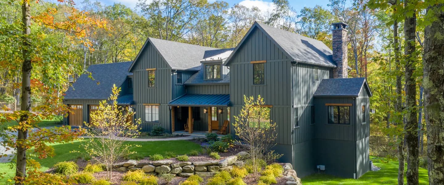 Dark grey board and batten farmhouse with gables, a timber porch and a stone chimney among autumn trees
